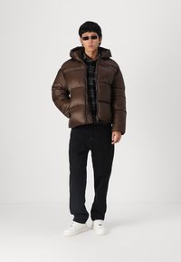 Chaqueta acolchada marrón con capucha, con un diseño de acolchado. Llevada sobre una camisa de cuadros negra y combinada con jeans negros y zapatillas blancas.