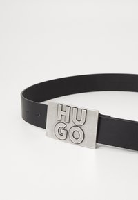 Črn usnjen pas z ovalno sponko srebrne barve, na kateri je vkovana beseda "HUGO". Gladka tekstura in minimalističen dizajn.