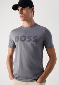 Camiseta gris de manga corta hecha de algodón suave, con un prominente logo en gris oscuro "BOSS" en el pecho. Diseño simple y casual.