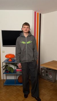 Ung mann i grå hettegenser og mørke bukser står i et rom med en veggmontert TV, oransje lampe, basketball og fargerike vertikale striper på hjørneveggen.