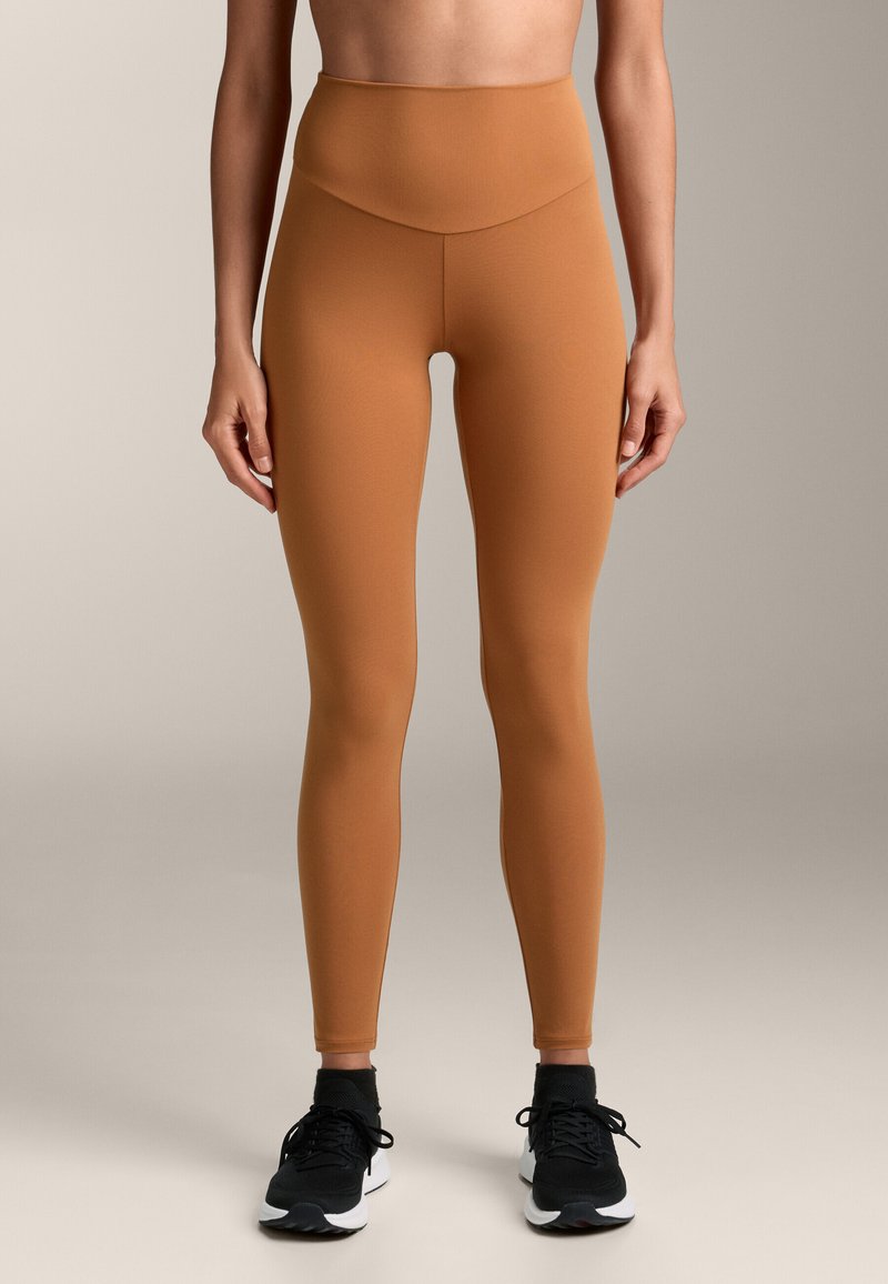 Rjave legice visoke pasu, narejene iz raztegljivega blaga, brez šivov, v kombinaciji s črnimi športnimi čevlji z belimi poudarki.