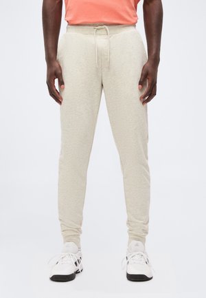 Man draagt lichtbeige joggerbroek met aantrekkoord, witte sportschoenen met zwarte zolen en een koraalroze T-shirt met korte mouwen tegen een effen achtergrond.