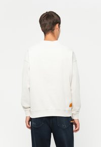 Sudadera blanca de corte relajado, de manga larga, con puños acanalados y un pequeño logotipo naranja en la parte inferior. Combinada con pantalones vaqueros oscuros.