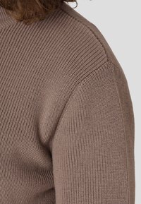 Nahaufnahme eines braunen Strickpullovers, der die gerippte Textur, einen Rundhalsausschnitt und eine markante Schulternaht zeigt.