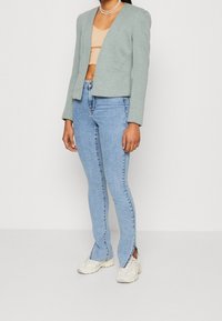 Blazer texturé vert clair à coupe courte, associé à un jean bleu moulant avec des fentes aux chevilles, et des baskets blanches.