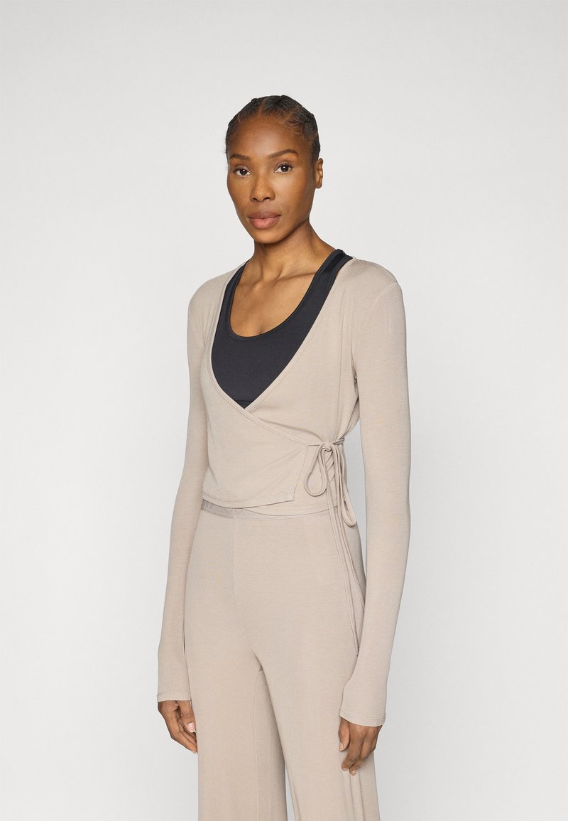 Beżowa bluzka kopertowa z długimi rękawami, ozdobiona sznurkiem w talii, noszona na ciemnej czarnej koszulce na ramiączkach. Gładka tekstura.