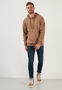 Sudadera con capucha marrón, de textura acanalada y detalles de costuras diagonales, combinada con jeans oscuros de corte ajustado y zapatillas altas en tono marrón claro.