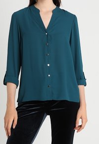 Tealfärgad blus med V-ringning och knappstängning, med uppvikbara ärmar och en mjuk tygstruktur. Silverknappar ger en detalj.