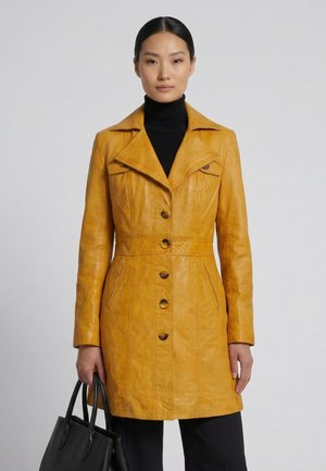 Kvinde i en sennepsgul knap-læderjakke, sort rullekrave, sorte bukser, der holder en sort håndtaske mod en ensfarvet lys baggrund.