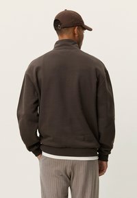 Καφέ φούτερ με φερμουάρ στο ένα τέταρτο, σε άνετη γραμμή, με ριμπ μανσέτες και τελείωμα. Φορεμένο πάνω από λευκό πουκάμισο και συνδυασμένο με παντελόνι με γκρι ρίγες.