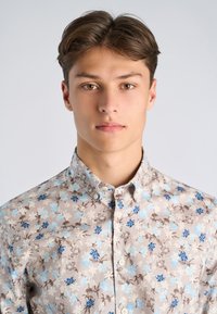 Noormees helepruunide juustega, kandes beeži värvi nööpidega särki siniste ja pruunide lillemustritega, näoga otsevaates lihtsa tausta ees.