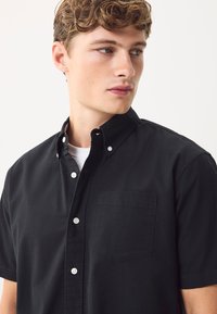 Camicia nera a maniche corte con bottoni, colletto appuntito, tasca sul petto a sinistra e bottoni bianchi. Il tessuto sembra liscio con una texture solida.