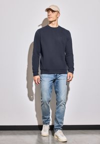 Navy blue sweatshirt with crew neckline and long sleeves, paired with light blue jeans and white sneakers. Model stands against a neutral backdrop.