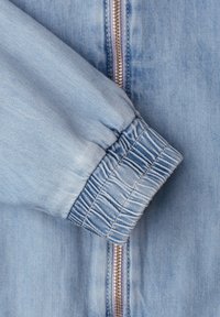 Light blue denim jacket sleeve with elastic cuff next to a closed brass zipper on the front body of the jacket.