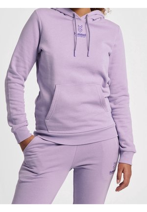 Person trägt hellvioletten Hoodie und passende Jogginghose mit der Hand in der Tasche und zeigt Details des entspannten lässigen Outfits sowie das Logo.