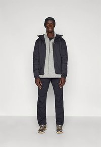 Uomo con berretto nero, giacca a vento nera sopra un maglione grigio con zip, pantaloni neri e scarpe sportive, in piedi contro uno sfondo bianco semplice.