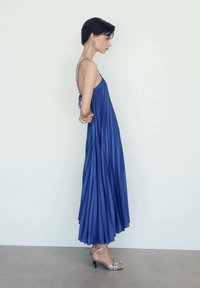 Modra maksi obleka z nabrusi in tankimi naramnicami, ki ima zavezovanje zadaj, v kombinaciji s srebrnimi sandalnimi petami. Gladek material in eleganten padec.