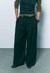 Črne široke hlače z gubami, izdelane iz gladkega materiala. V kombinaciji z temno zelenim rebrastim puloverjem in črnimi balerinkami s odprtim prstom.