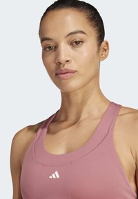 Mujer con el cabello oscuro recogido, vestida con un sujetador deportivo rosa de Adidas, mirando al frente con una expresión neutral sobre un fondo claro.