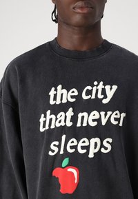 Black sweatshirt with ribbed neckline, featuring white text "the city that never sleeps" and a red apple graphic with a green leaf.