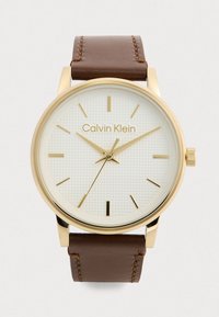 Montre Calvin Klein avec un boîtier en acier inoxydable doré, un bracelet en cuir brun, un cadran blanc texturé, des index horaires dorés et un design minimaliste.