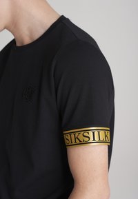 Camiseta negra con un diseño ajustado. Presenta una banda de brazo a rayas doradas con la inscripción "SIKSILK" en letras negritas. Textura de algodón suave.