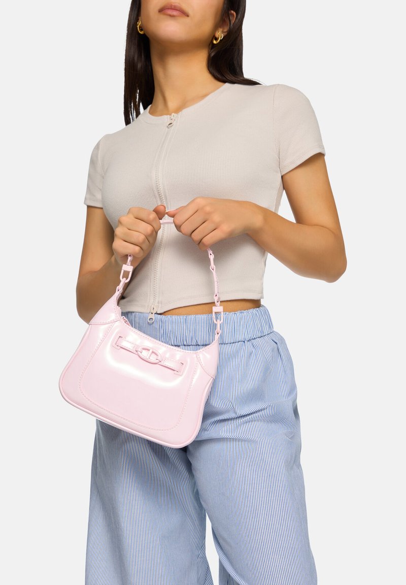 Liten handväska i rosa lackläder med platt form och kort handtag som har en rosettdekoration. Modell som bär en åtsittande beige topp och randiga byxor.