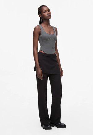 Femme aux cheveux tressés portant un débardeur gris ajusté, une superposition de jupe noire, un pantalon large noir et des chaussures noires épaisses, debout devant un fond blanc.