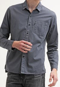 Camisa de manga larga gris con botón, cuello punta, bolsillo en el pecho y textura sutil; botones en la parte delantera; silueta ajustada.