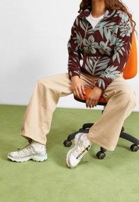 Polarowy sweter z kwiecistym wzorem w brązie i teal, beżowe spodnie w szerokim kroju oraz srebrne buty sportowe z akcentami. Siedzi na krześle.