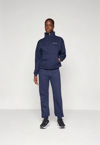 Tengerészkék pulóver magas nyakkal és cipzárral, hozzáillő nadrággal. Az anyaga puha, texturált polár. Márkás logóval díszítve.