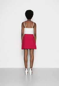 Valkoinen pitsireunainen toppi, joka on yhdistetty punaiseen aaltoilevahelmaiseen hameeseen. Mallilla on päällään valkoiset korot, ja hänen sileät jalkansa näkyvät yksinkertaisella taustalla.