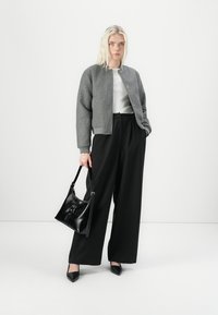 Giacca bomber in lana grigia sopra una maglia bianca, abbinata a pantaloni neri a gamba larga. Tiene una borsa in pelle nera e indossa tacchi a punta neri.