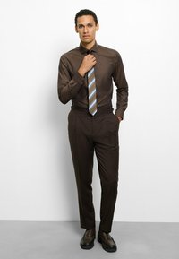 Braunes Hemd mit feiner Struktur, kombiniert mit passenden braunen Hosen. Krawatte mit Streifen in Braun, Blau und Weiß. Schwarze Schuhe mit dicker Sohle.