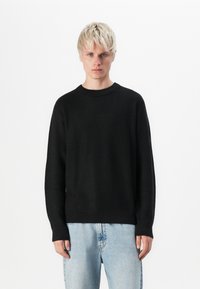 Pull noir avec un motif de tricot texturé, col à côtes et poignets, associé à un jean bleu clair. Design simple et classique.