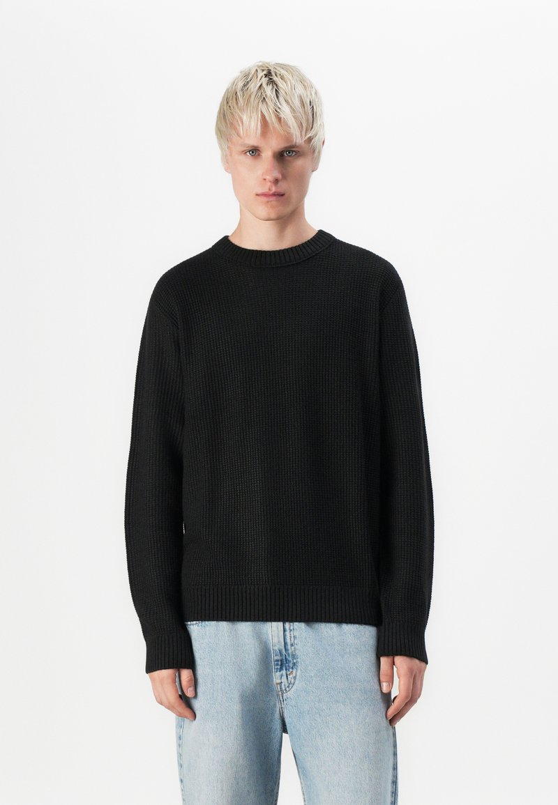 Pull noir avec un motif de tricot texturé, col à côtes et poignets, associé à un jean bleu clair. Design simple et classique.