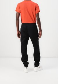 Svarta byxor med elastisk midja och två bakfickor, ihop med en orange t-shirt och vita sneakers, sedd från ryggen.