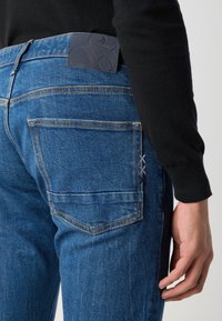 Jeans de mezclilla azul con una etiqueta de cuero negro texturizado en la cintura, que cuentan con cinco bolsillos y detalles de costuras visibles en naranja y púrpura.