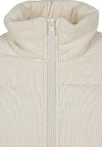 Krämfärgad, ribbad pufferjacka med hög krage, framknäppning med dragkedja och mjuk textur. Anmärkningsvärd sömdetaljering på kroppen.