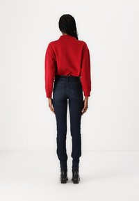 Κόκκινη φούτερ μπλούζα με γιακά, συνδυασμένη με σκούρα μπλε ψηλόμεσα τζιν. Χαρακτηρίζει εφαρμογή στενής γραμμής, λεία υφή και μονόχρωμα σχέδια. Μαύρες ψηλές μπότες.