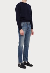 Henkilö, jolla on päällä tummansininen neule, vaaleansiniset slim-fit-farkut, valkoiset sukat ja mustat nahka-loaferit, seisoo yksinkertaista valkoista taustaa vasten.