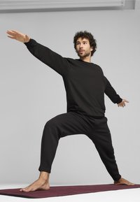 Schwarzer sportlicher Sweatshirt und strukturierte schwarze Hose, während einer Yoga-Pose auf einer kastanienbraunen Matte, barfuß und mit ausgestreckten Armen.