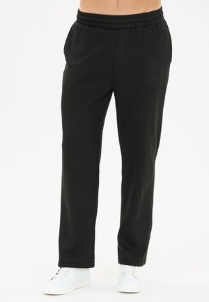 Pantalon de survêtement noir à taille élastique porté avec des baskets blanches sur une personne debout, visible de la taille aux pieds sur fond blanc.