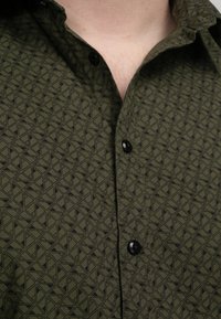 Camisa de color verde oscuro con un patrón geométrico, botones negros y un cuello estructurado. La tela tiene una textura suave.