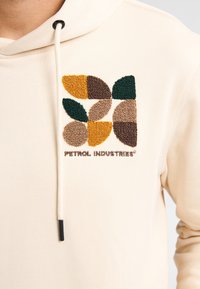 Sudadera con capucha color beige que presenta un gráfico bordado con hojas en verde, amarillo, marrón y beige, y el texto "PETROL INDUSTRIES".