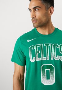 Green cotton t-shirt featuring "CELTICS" in white with a gradient effect and a prominent "0" below. Nike logo on the left shoulder.