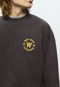 Osoba s crvenkastom kosom koja nosi tamnosmeđi duksericu s žutim logotipom "Wood Wood Copenhagen 2002" na prsima.