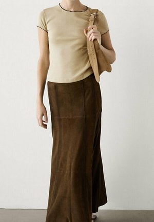 Femme portant un haut beige à manches courtes avec un liseré noir et une longue jupe en daim marron, tenant un sac à bandoulière en daim marron assorti.