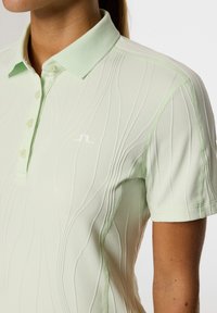 Hellgrünes Poloshirt mit strukturiertem Design, das dezente Wellenmuster zeigt, einem Kragen und einem kleinen weißen Logo auf der Brust.