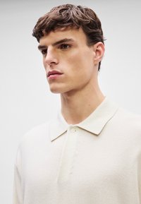 Λευκή πόλο μπλούζα από μαλακό ύφασμα, με κλασικό γιακά, κλείσιμο με ένα κουμπί και διακριτή υφή. Ουδέτερο φόντο.